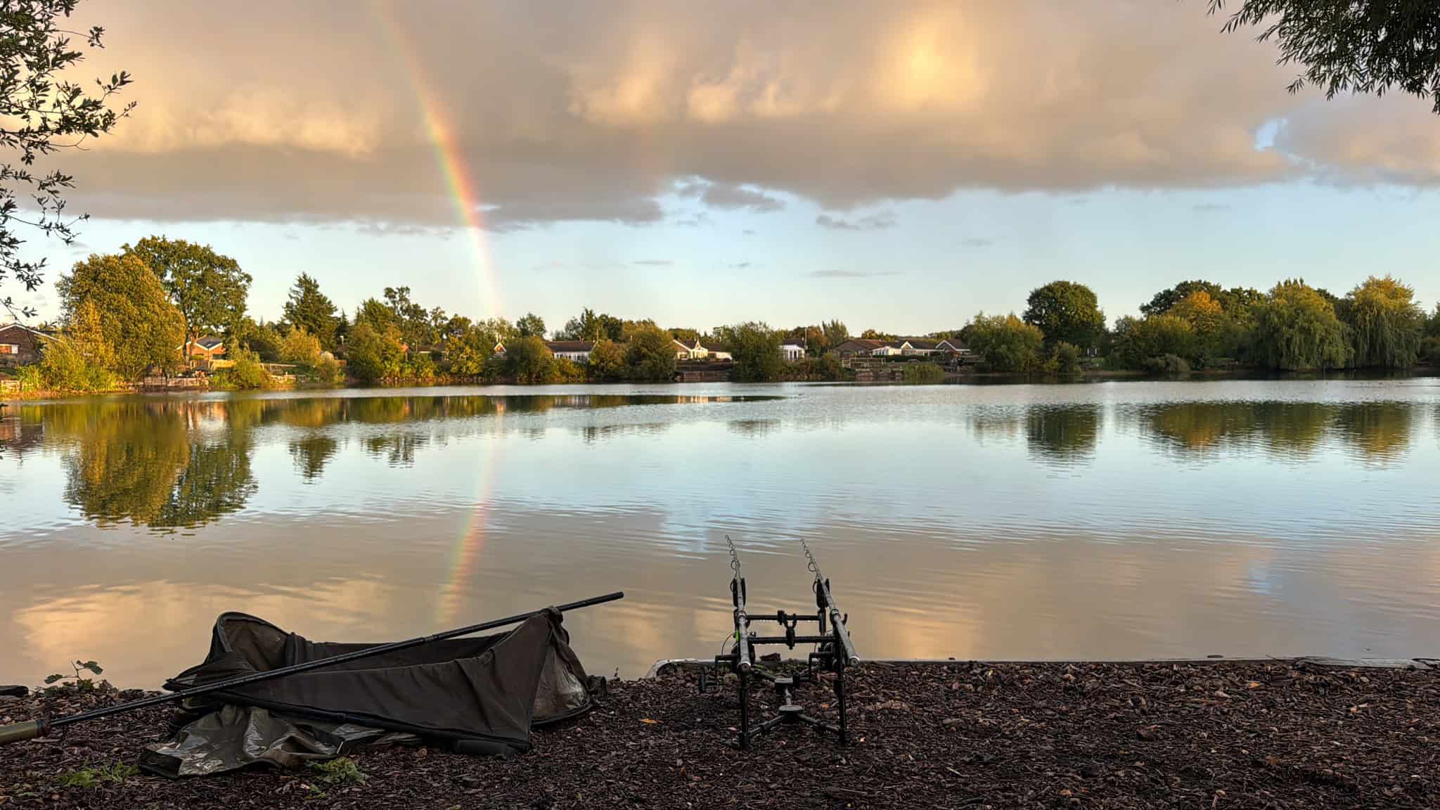 Rainbow over peaceful lake with fishing gear in foreground, serene outdoor fishing scene for Carp Tax SEO.